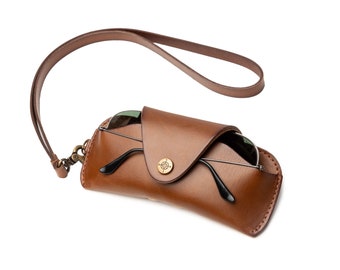 May include: A brown leather glasses case with a strap. The case has a flap that closes with a brass button. The case is empty and the strap is hanging loose.
