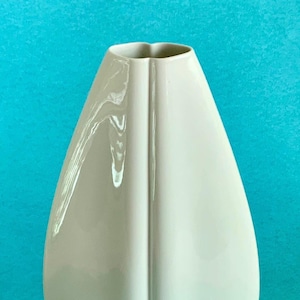 Könnte beinhalten: Eine hohe, cremefarbene Keramikvase mit einer einzigartigen, länglichen Form. Die Vase hat eine glatte, glänzende Oberfläche und eine vertikale Rille in der Mitte, die einen Hauch von Eleganz verleiht. Die Vase ist ein dekorativer Gegenstand.
