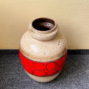 Könnte beinhalten: Eine Keramikvase mit rundem Körper und schmalem Hals. Die Vase hat eine gesprenkelte beige Oberfläche mit einem Band aus roten Kreisen in der Mitte. Der Innenraum ist dunkelbraun.