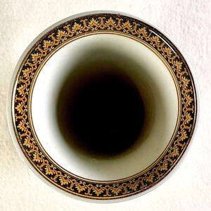 May include: A white ceramic vase with a black interior. The rim features a decorative border with gold and black floral patterns. The vase has a smooth, glossy finish and a classic design.