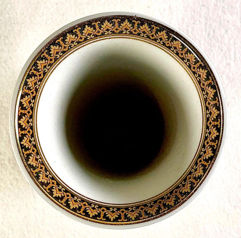 May include: A white ceramic vase with a black interior. The rim features a decorative border with gold and black floral patterns. The vase has a smooth, glossy finish and a classic design.