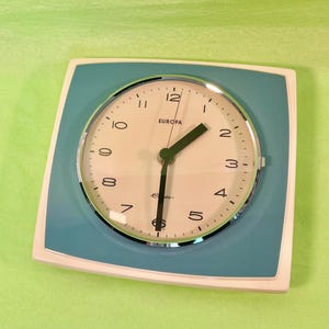 Könnte beinhalten: Eine quadratische Wanduhr mit cremefarbenem Zifferblatt und türkisfarbenem Rand. Die Uhr hat schwarze Ziffern, einen chromfarbenen runden Rahmen und schwarze Zeiger. Das Wort EUROPA ist schwarz über der Mitte aufgedruckt, darunter steht Electronic.