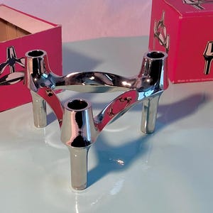 Può includere: Un portacandele argentato lucido con tre aperture cilindriche e tre gambe. Il design presenta una struttura curva e intrecciata. Il portacandele è esposto su una superficie azzurra, con una scatola rosa sullo sfondo.