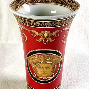 May include: Red and gold decorative vase with a Medusa head emblem. The vase features ornate gold detailing and a black border. The top and bottom rims have a black and gold pattern. A luxury home decor item.