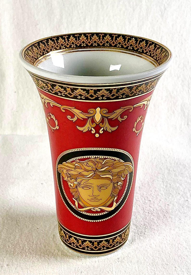 May include: Red and gold decorative vase with a Medusa head emblem. The vase features ornate gold detailing and a black border. The top and bottom rims have a black and gold pattern. A luxury home decor item.