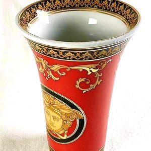 May include: A red ceramic vase with gold accents and a Medusa head design. The vase has a wide opening and a tapered shape. The rim and base feature intricate gold patterns. The vase is approximately 8 inches tall.