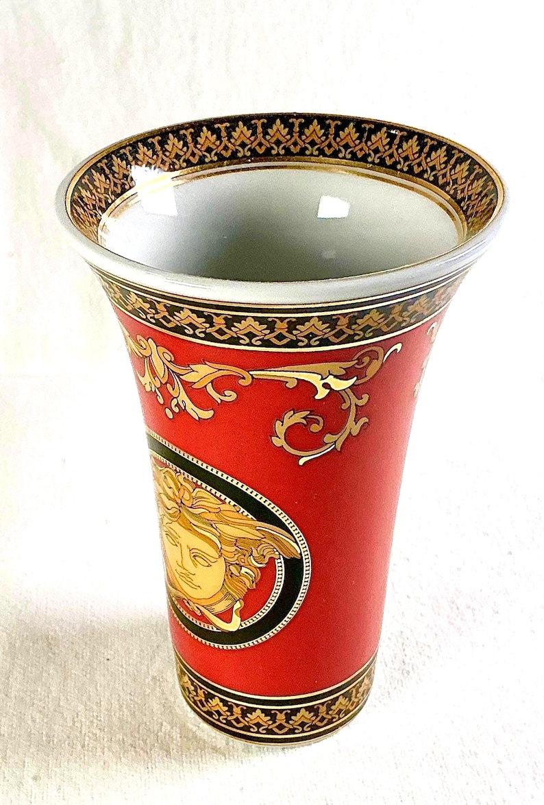 May include: A red ceramic vase with gold accents and a Medusa head design. The vase has a wide opening and a tapered shape. The rim and base feature intricate gold patterns. The vase is approximately 8 inches tall.
