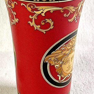 May include: A red vase with gold and black decorative accents. The vase features a gold Medusa head emblem and ornate gold scrollwork. The top and bottom rims have a black and gold patterned border.