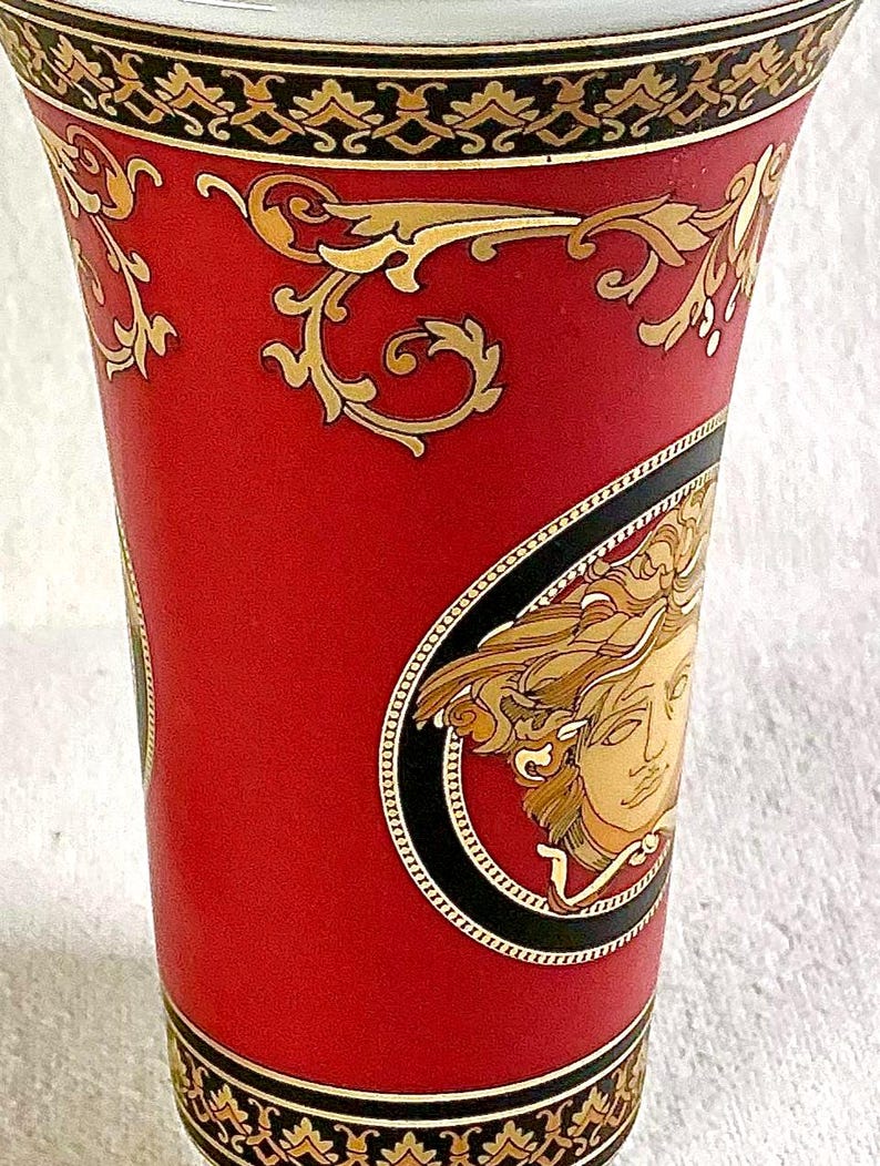May include: A red vase with gold and black decorative accents. The vase features a gold Medusa head emblem and ornate gold scrollwork. The top and bottom rims have a black and gold patterned border.