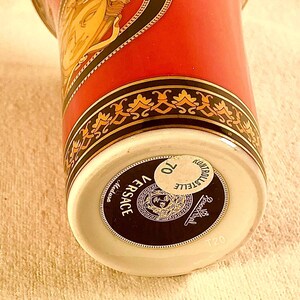 May include: A close-up of a Versace Medusa ceramic cup. The cup features a red background with gold and black decorative accents. The Versace logo is prominently displayed on the bottom of the cup.