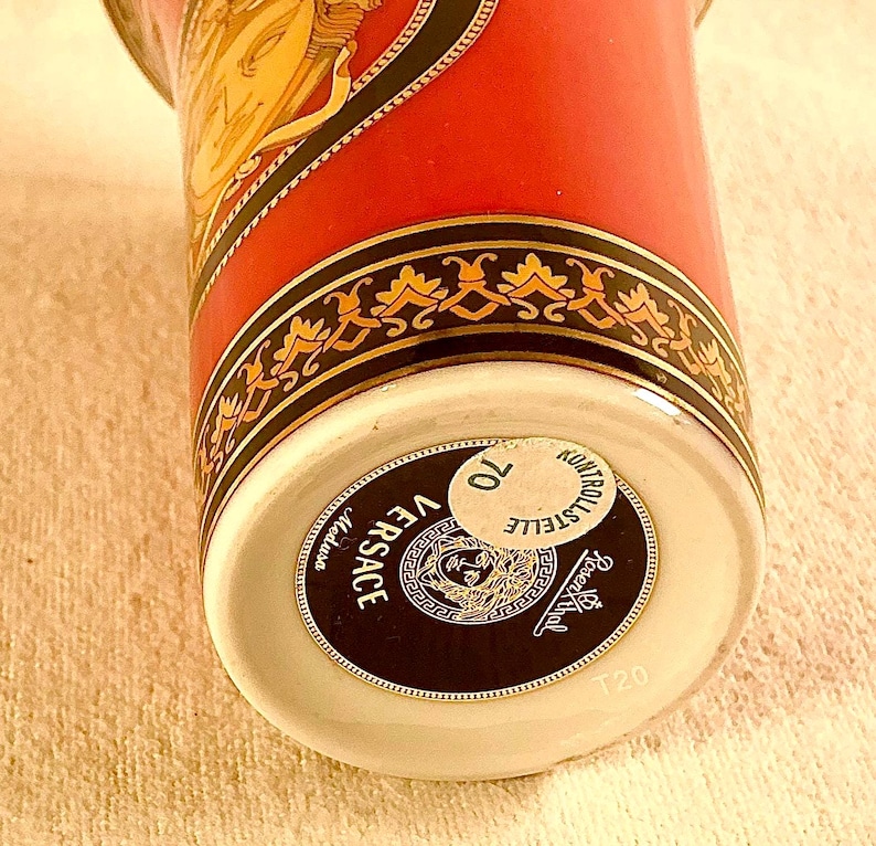 May include: A close-up of a Versace Medusa ceramic cup. The cup features a red background with gold and black decorative accents. The Versace logo is prominently displayed on the bottom of the cup.