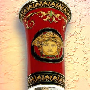 May include: A red ceramic vase with gold and black decorative trim. The vase features a gold Medusa head emblem on the front. The top and bottom rims have intricate gold patterns. The vase has a white base.