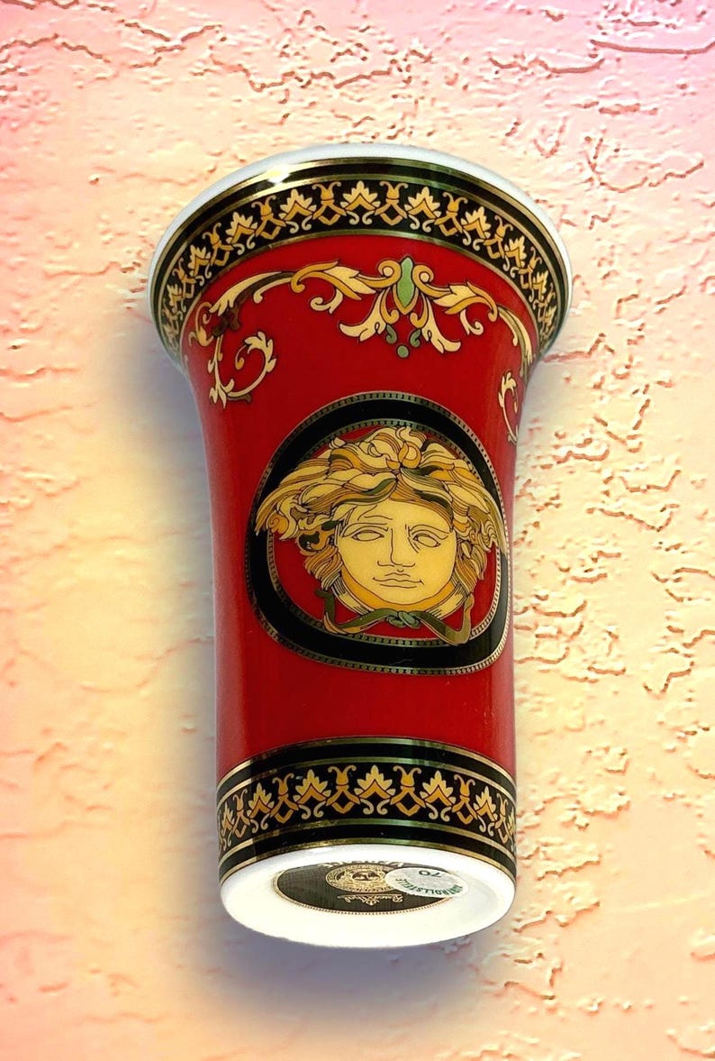 May include: A red ceramic vase with gold and black decorative trim. The vase features a gold Medusa head emblem on the front. The top and bottom rims have intricate gold patterns. The vase has a white base.