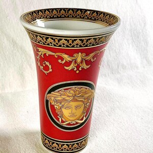 May include: Red ceramic vase with gold and black decorative accents. Features a gold Medusa head emblem within a black oval frame. The rim and base have intricate gold and black patterns. A luxury home decor item.