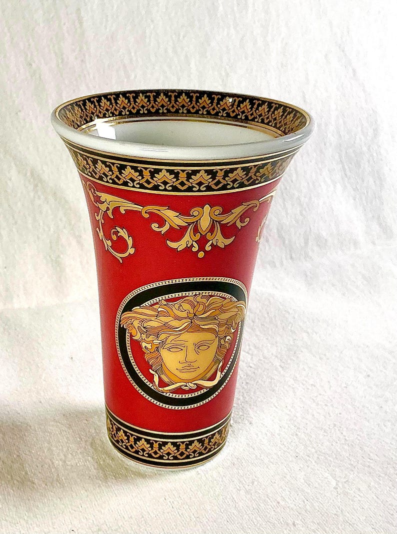May include: Red ceramic vase with gold and black decorative accents. Features a gold Medusa head emblem within a black oval frame. The rim and base have intricate gold and black patterns. A luxury home decor item.