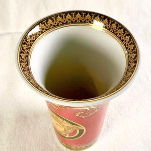 May include: A decorative vase with a wide, gold-trimmed rim featuring an ornate black and gold pattern. The vase has a red body with gold accents and a classical design. The interior is white.
