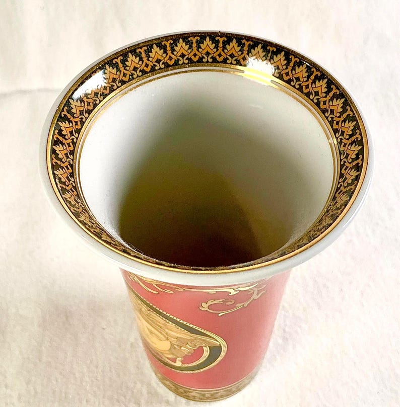 May include: A decorative vase with a wide, gold-trimmed rim featuring an ornate black and gold pattern. The vase has a red body with gold accents and a classical design. The interior is white.