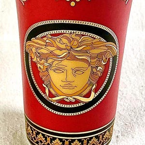 May include: A red ceramic vase with gold accents and a central oval medallion featuring a gold Medusa head design. The vase has a black and gold border at the top and bottom.