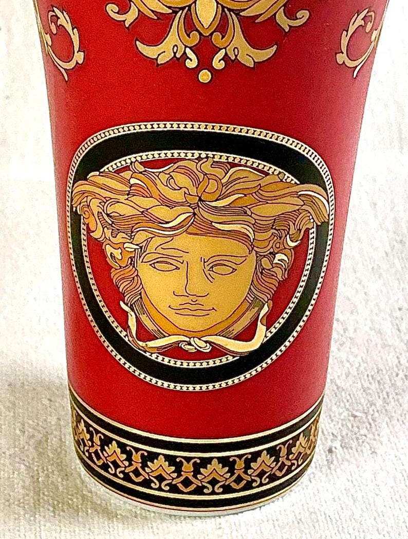May include: A red ceramic vase with gold accents and a central oval medallion featuring a gold Medusa head design. The vase has a black and gold border at the top and bottom.