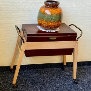 Könnte beinhalten: Ein Nähwagen aus Holz mit einem dunkelbraunen Deckelfach, hellen Holzbeinen und Metallgriffen an beiden Seiten. Eine runde Keramikvase mit gesprenkelter orange-grüner Glasur steht auf einer kleinen weißen Spitzendecke auf dem Wagen.