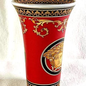 May include: A red vase with gold and black decorative patterns. The vase features a Medusa head emblem and ornate gold scrollwork. The rim and base have black and gold trim. The vase is a decorative home decor item.