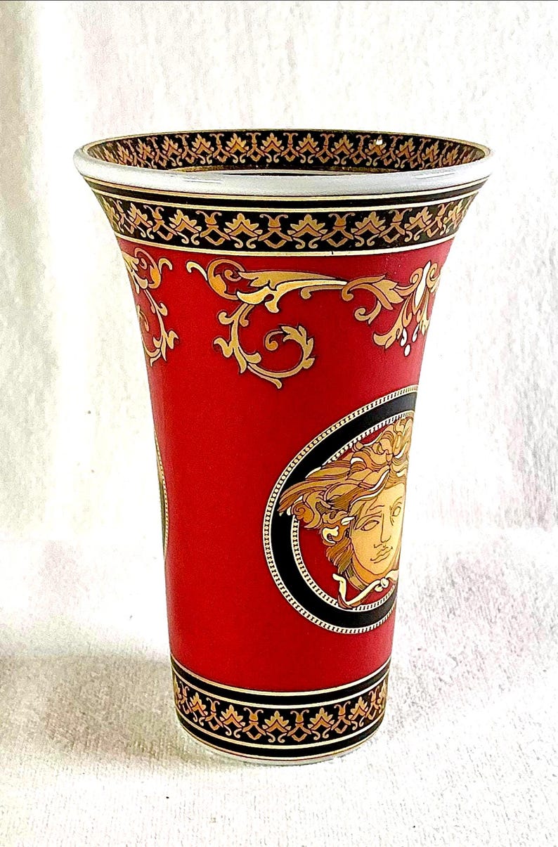May include: A red vase with gold and black decorative patterns. The vase features a Medusa head emblem and ornate gold scrollwork. The rim and base have black and gold trim. The vase is a decorative home decor item.