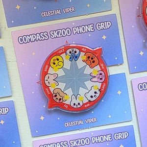 Peut inclure: Plusieurs supports de téléphone rouges avec un design de boussole, des visages d'animaux de dessin animé et le texte "COMPASS SKZOO PHONE GRIP". Ils sont présentés sur des cartes avec un fond dégradé violet et bleu et le texte "CELESTIAL VIPER".
