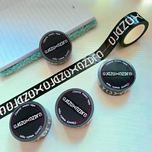 Może przedstawiać: Czarna taśma washi z białym tekstem "OJAZUXOZDRO" i "Clancy washi tape". Taśma jest rozwinięta i pojawia się również w rolkach. Taśma ma czerwony pasek pośrodku. Rolki są szare i czarne.