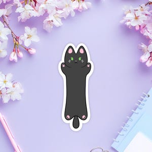 Peut inclure: Un autocollant de chat noir avec des yeux verts et des pattes roses. Le chat est dans une pose allongée, les bras levés.