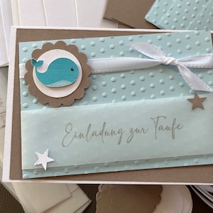May include: A light blue card with white polka dots and a white ribbon. The card has a brown scalloped circle with a blue whale illustration and the text "Einladung zur Taufe".