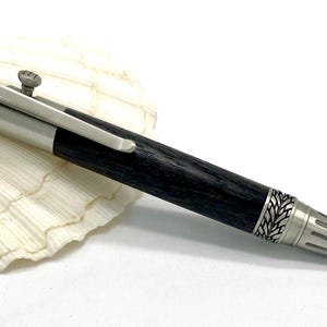 May include: A handcrafted pen with a black wooden barrel and silver-tone metal accents. The pen features intricate leaf-like designs on the ends and a clip. The pen is resting on a white seashell.