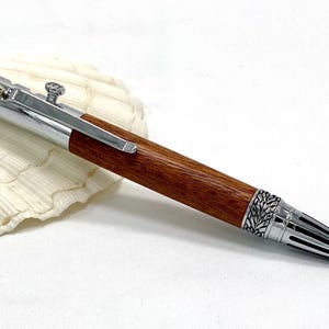 May include: A handcrafted pen with a wooden barrel and silver accents. The pen has a decorative silver cap and tip with a leaf design. The pen is resting on a white seashell.