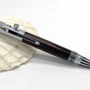 May include: A handcrafted pen with a dark wood barrel and silver-colored metal accents. The pen features intricate detailing on the tip and end, and a small knob on the side. The pen is resting on a white seashell.