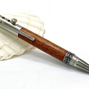 Könnte beinhalten: Handgefertigter Stift mit Holzschaft und silberfarbenen Metallakzenten. Der Stift hat ein detailliertes Blattdesign an Spitze und Ende, mit einer strukturierten Oberseite. Der Stift wird auf einer weißen Muschel präsentiert.