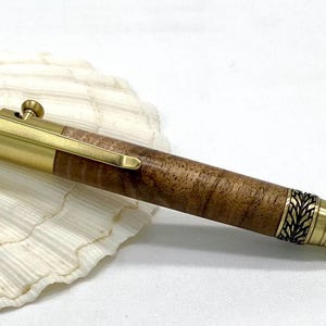 Könnte beinhalten: Ein handgefertigter Stift mit Messing-Finish und Holzschaft. Der Stift hat aufwendige blattartige Details an den Enden und einen Clip. Der Stift liegt auf einer weißen Muschelschale.