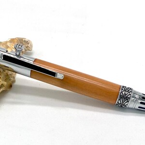 Peut inclure: Un stylo artisanal avec un corps en bois brun clair et des accents métalliques argentés. Le stylo présente des motifs de feuilles et géométriques complexes sur les composants métalliques. Le stylo est présenté sur une surface blanche à côté d'un coquillage.