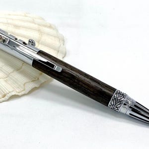 Könnte beinhalten: Ein handgefertigter Stift mit einem dunklen Holzschaft und silberfarbenen Metallakzenten. Der Stift hat aufwendige Details an der Spitze und am oberen Ende sowie einen silbernen Clip. Der Stift wird auf einer weißen Muschel präsentiert.