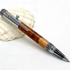 Op de afbeelding: Een handgemaakte pen met een zilverkleurige metalen behuizing en een houten vat. De loop heeft een natuurlijk houtnerfpatroon in bruin- en beigetinten. De pen heeft een decoratieve clip en een getextureerd uiteinde.