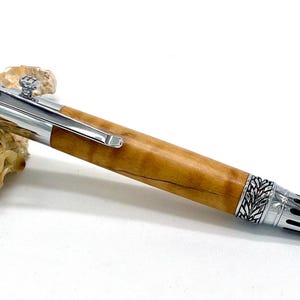 May include: A handcrafted pen with a light brown wooden barrel and silver-colored metal accents. The pen features intricate detailing on the tip and end, with a clip for securing it in a pocket. The pen is displayed on a white surface.