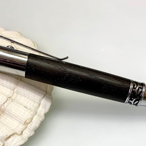 Könnte beinhalten: Ein schwarzer und silberner Stift mit dekorativem Design. Der Stift hat einen dunklen Holzschaft und einen silbernen Clip. Der Stift liegt auf einer weißen Muschelschale.