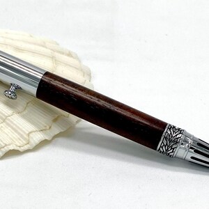 May include: A handcrafted pen with a dark brown wooden barrel and silver accents. The pen features intricate leaf-like designs on the silver components and a small button. The pen is resting on a white seashell.