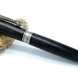 Peut inclure: Un stylo noir et argent avec un clip en forme de serpent et une pointe à motif diamant. Le stylo repose sur une coquille.