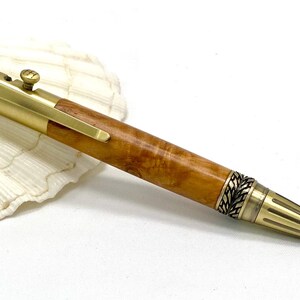 May include: A handcrafted pen with a wooden barrel and gold-coloured metal accents. The pen features a decorative leaf pattern on the ends and a clip. The pen is resting on a white seashell.
