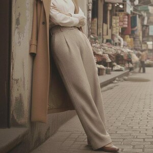 May include: A woman wearing a tan overcoat, white blouse, and light brown pants, leaning against a wall. The outfit is completed with brown flats. The scene is set on a brick-paved street with a blurred background of shops.