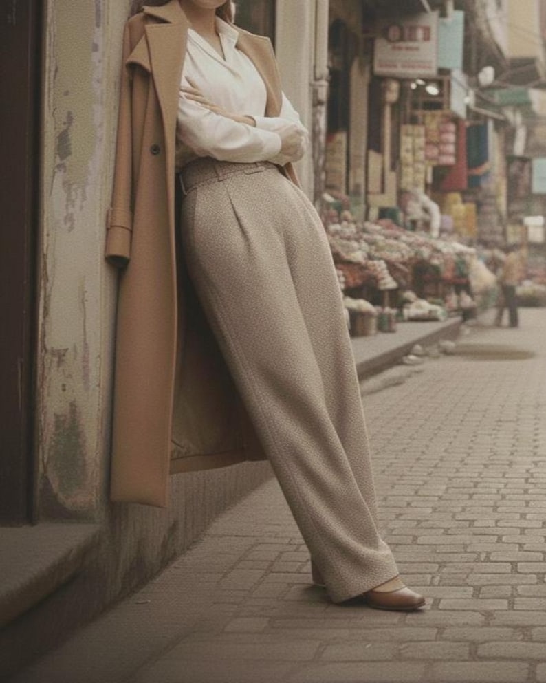 May include: A woman wearing a tan overcoat, white blouse, and light brown pants, leaning against a wall. The outfit is completed with brown flats. The scene is set on a brick-paved street with a blurred background of shops.