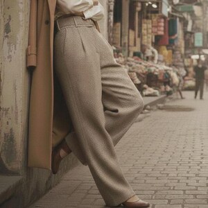 May include: A person wearing a tan overcoat, white blouse, and light brown trousers. The person is leaning against a wall. Brown leather flats complete the outfit. The background shows a street scene.