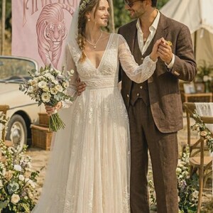 Puede incluir: Una pareja en una boda al aire libre. La novia lleva un vestido de novia de encaje blanco con mangas largas y una corona de flores. El novio lleva un traje marr&oacute;n y sombrero. Una pancarta con "PINK TIGER" y un tigre en el fondo.