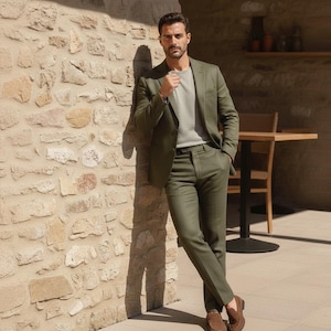 Op de afbeelding: Een man in een groen pak, een lichtgrijs crewneck shirt en bruine suède loafers. Het pak bestaat uit een blazer en bijpassende broek. De man leunt tegen een stenen muur. De setting lijkt buiten te zijn.