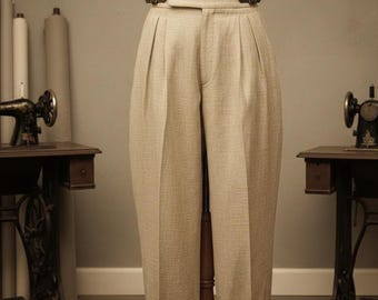 May include: Beige high-waisted trousers with a wide waistband and two brown leather buckle closures. The trousers have pleats at the front and are displayed on a mannequin. The cuffs are turned up with a white trim. Sewing machines are in the background.