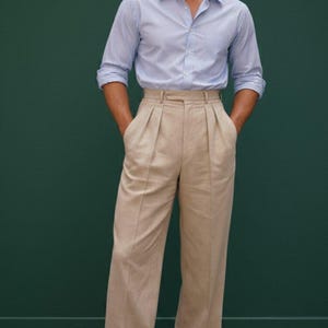 May include: A light blue button-down shirt with rolled-up sleeves and beige pleated trousers. The outfit is completed with brown leather dress shoes. The background is a solid dark green.